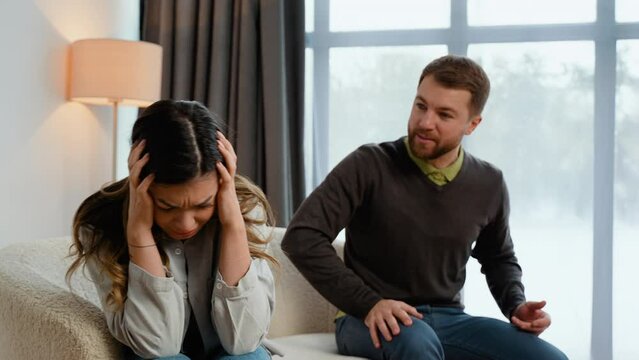 Caucasian couple sitting on sofa in living room, quarreling over disagreement, man yelling at woman, woman experiencing psychological abuse from her husband