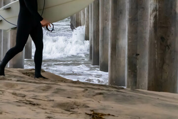 Unknown Surfer Holding Board Walking on Beach in Front of Underside of Pier with Crashing Waves in...