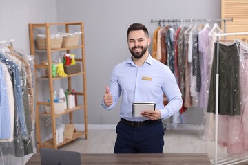 Dry-cleaning service. Happy worker with notebook showing thumb up in workplace indoors