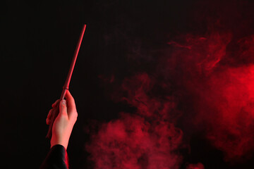 Magician holding wand in smoke on dark background, closeup. Space for text - obrazy, fototapety, plakaty