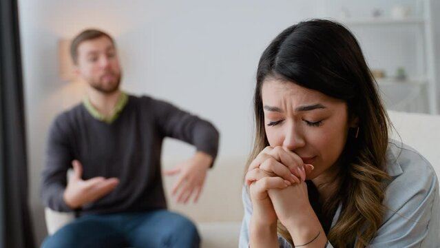 Tired stressed woman sadness looking away sitting on bed on blurred background of aggressive husband shouting on wife at home. Concept of family scandal, crisis, domestic violence, abuse