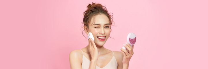 Beautiful young woman applying face powder with puff applicator on pink background