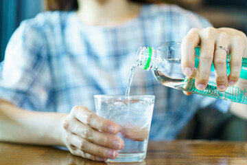 Unrecognizable woman pouring drinking water from a plastic bottle into drinking glass on wooden table close up.
