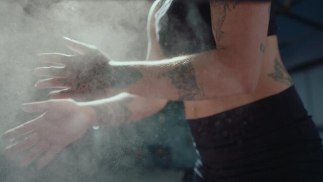 Muscular woman in sports shorts and bra covering palms with chalk powder before weightlifting workout in a gym. Slow mo, tilt-up shot