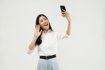 Photo of a young Asian woman wearing a white shirt, taking a selfie on a smartphone, video chatting with a mobile phone app, and isolated on a white background.