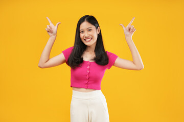 A portrait of a young Asian woman with a satisfied expression, points away with both forefingers up for a copy space, looking at the camera, isolated on a yellow background. - obrazy, fototapety, plakaty