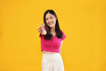 Portrait of a young Asian woman with a pink t-shirt showing a korean heart isolated on a yellow background, fall in love concept.