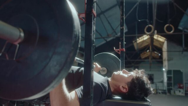 Young strong man doing barbell bench press: he lying down on a flat bench and pressing weighted barbell up and down during training in the gym