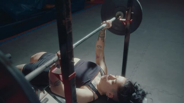 Young female athlete with muscular tattooed body performing barbell bench press when training in the gym. Tilt-down shot, slow mo