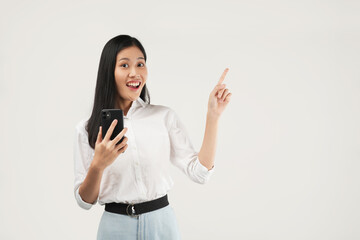 Photo of young asian girl shopping on mobile app, holding phone and pointing finger left at empty copy space, showing promo text, standing with smartphone against white background