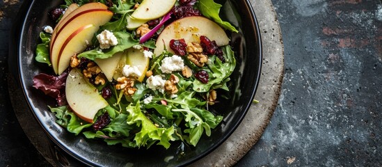 Above view of a black bowl on a concrete table with a green salad featuring apples, goat cheese, cranberries, red onion, and pepitas.