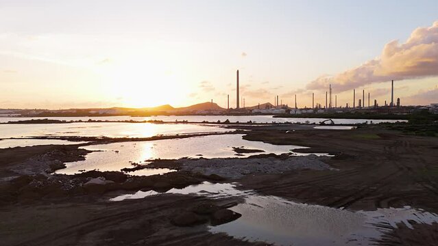 Aerial dolly above asphalt lake and white piles of refuse at sunset with oil refinery stacks in distance
