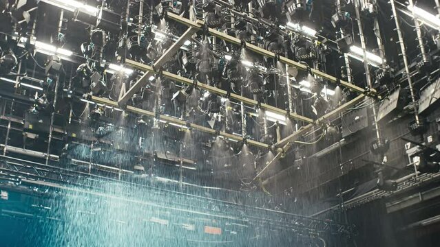 A special effects rain machine in a film cinema studio with a ceiling full of film lights. Wide shot of the full device with wooden frame, hoses and sprinklers. Music video strobe lights are flashing.