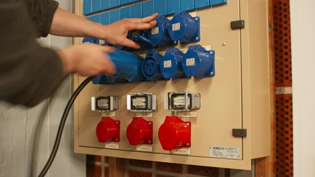 Electrical panel with blue 16A and red 32A three phase connectors and distribution box. Technician unplugging and removing three cables from adjacent sockets.