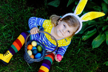 Cute little toddler girl with bunny ears having fun with traditional Easter eggs hunt on warm sunny...