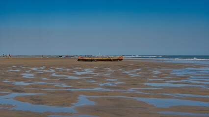 Low tide in the ocean. Traditional Malagasy wooden pirogue boats are located on the exposed seabed. Tiny silhouettes of people, a sailboat in the distance.The blue sky is reflected in puddles of water - obrazy, fototapety, plakaty