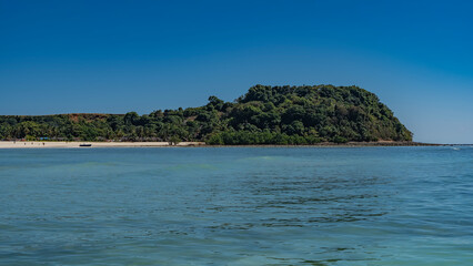 The beautiful tropical island  with lush green vegetation. Tiny silhouettes of people, moored boats, and houses of local residents are visible on the sandy beach. Clear blue sky, turquoise ocean. 