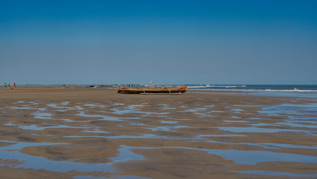 Low tide in the ocean. Traditional Malagasy wooden pirogue boats are located on the exposed seabed. Tiny silhouettes of people, a sailboat in the distance.The blue sky is reflected in puddles of water