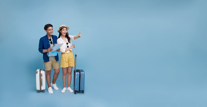 Happy Asian couple tourist looking map and hand pointing to copy space with baggage going to travel on holidays isolated on blue panorama background.