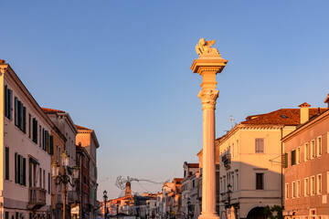 Picturesque Colonna Vigo, a column with majestic lion statue, illuminated by first rays of morning sun. Vibrant and charming Corso del Popolo street of Chioggia, Venetian Lagoon, Veneto, Italy