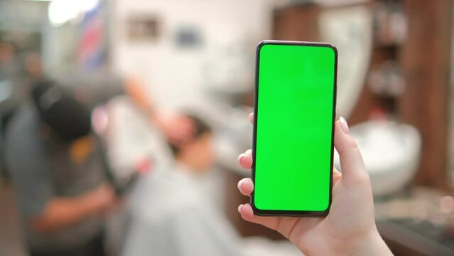 Barbershop. Back view of brunette holding chroma key green screen smartphone watching content. Gadgets and contemporary people concept. 4K