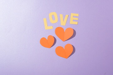 The word LOVE and red paper hearts are displayed on a pastel purple background. Space with a flat top-down perspective. Valentine's Day theme for advertising.