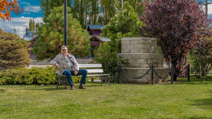 Naklejka na ściany i meble An adult man in a down jacket is relaxing while sitting on a park bench. Green grass on the lawn. Autumn. The leaves on the trees turned red. Cottages in the distance. Blue sky, clouds. Argentina.
