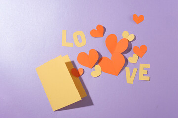 Flat lay of a card, the word LOVE and paper hearts arranged on a pastel purple background. Empty space for anniversary text design. View from above.