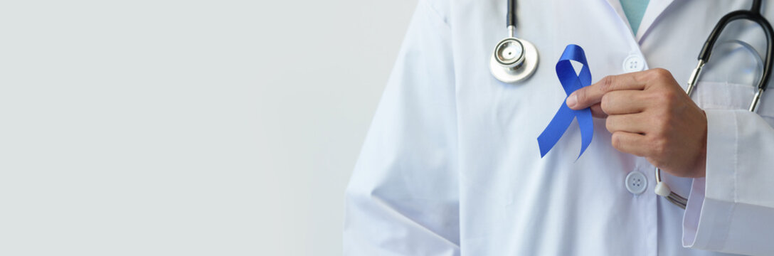 An image of a woman's hand in a doctor's uniform and stethoscope holding a blue ribbon represents fighting for life. Treatment from colon cancer World cancer day concept, copy space, banner, panorama