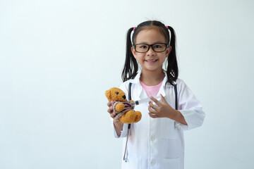Smiling girl wearing doctor's coat plays with her toy bear in medical practice Using a stethoscope and syringe for medical examination standing on a white background.