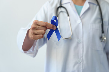 Image of a woman's hand in a doctor's uniform with a stethoscope holding a blue ribbon representing fighting for life saving lives and suffering from colon cancer. World cancer day concept, insurance