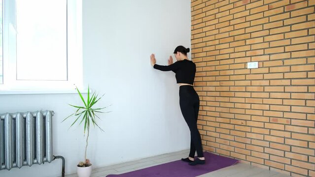 Slim sporty 30s woman lean her hand on the wall and rising leg. Workout at home. Having an active morning. Strengthening muscles, health concept. Healthy body practicing. Girl in black fitness suit