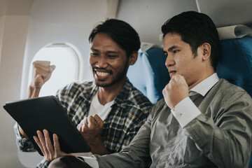 two Asian men on an airplane, looking at a tablet together, possibly friends choosing a location or...