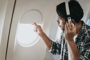 Asian man on an airplane looking out the window with headphones on, possibly enjoying music or an...