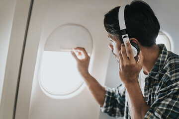 Asian man on an airplane looking out the window with headphones on, possibly enjoying music or an...