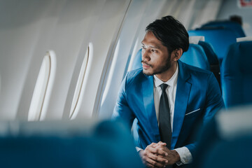 Asian businessman working on his laptop while seated in an airplane, smiling, possibly engaged in work or leisure during his flight.