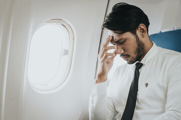 Asian businessman in a white shirt and tie seated in an airplane, with a hand on his temple...