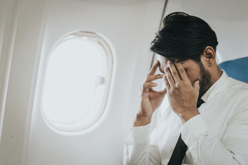 Asian businessman in a white shirt and tie seated in an airplane, with a hand on his temple...