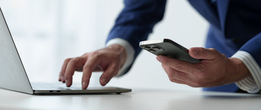 Businessman Holds Smartphone, Checks Email, Checks Job Details On Laptop Computer On White Desk To Point To Successful Business Goals In Office.