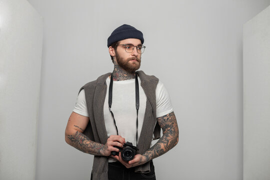Fashion handsome man photographer hipster with a beard with glasses with a tattoo in fashionable clothes with a knitted cap and a sweater holds a mirrorless photo camera in the studio