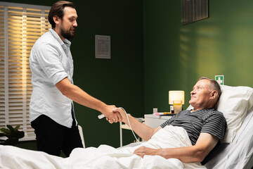 Son visits dad in hospital. They greet each other with a handshake. The older man is happy to see...