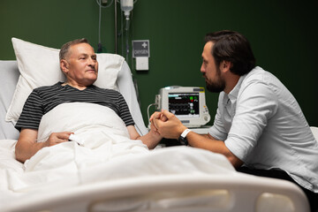 Obraz premium An elderly man lies in a hospital bed. A son visits his dad in the hospital. He holds his hand giving him support in difficult times