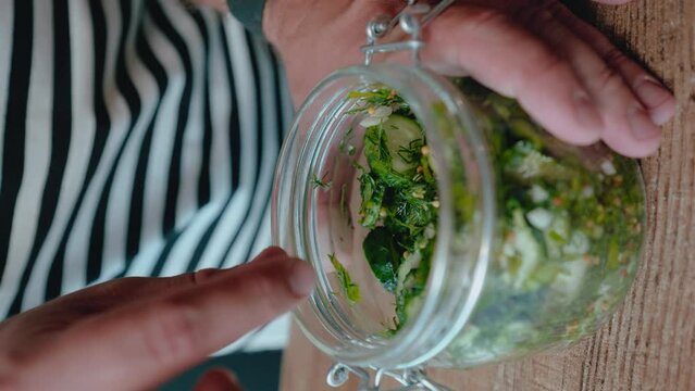 person in striped shirt is placing cucumber slices with herbs into glass jar on wooden surface. image focuses on hand and jar capturing the process of preparing homemade pickles or marinated cucumbers