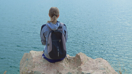 A woman tourist with a backpack on her shoulders sits on the top of a mountain and looks at the ocean.