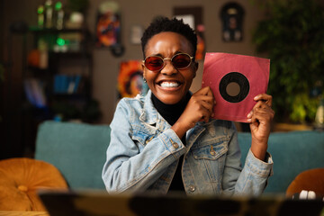 Portrait of dark skinned young woman in pink sunglasses smiling holding cd in hands showing at...