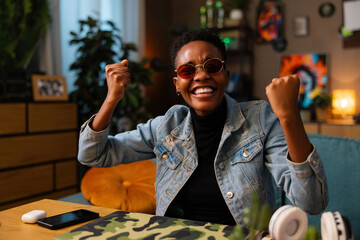 Happy african american teen girl lady woman with short hair using cell phone holding in hands smiling celebrating raising fists.