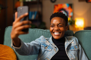 Portrait of delighted afo american dark skinned woman blogger vlogger taking selfie photo on camera sitting at home at weekends in cozy living room making stream online live. - obrazy, fototapety, plakaty