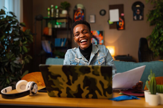 Delighted afro american dark skinned woman working online sitting in front of the laptop smiling observing papers work online from distance freelancer concept. - Powered by Adobe