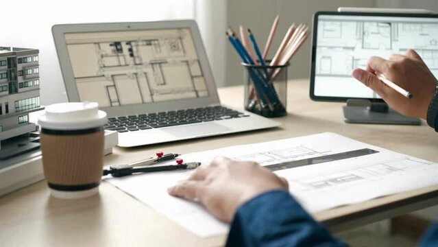 Professional Architect holding pencil working with laptop and blueprints for architectural plan. Closeup of engineer hand drawing on building blueprint with architectural equipment.