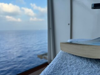 Buch liegt auf Stuhl und Handtuch einer Balkonkabine eines Kreuzfahrtschiffes mit Ausblick aufs Meer